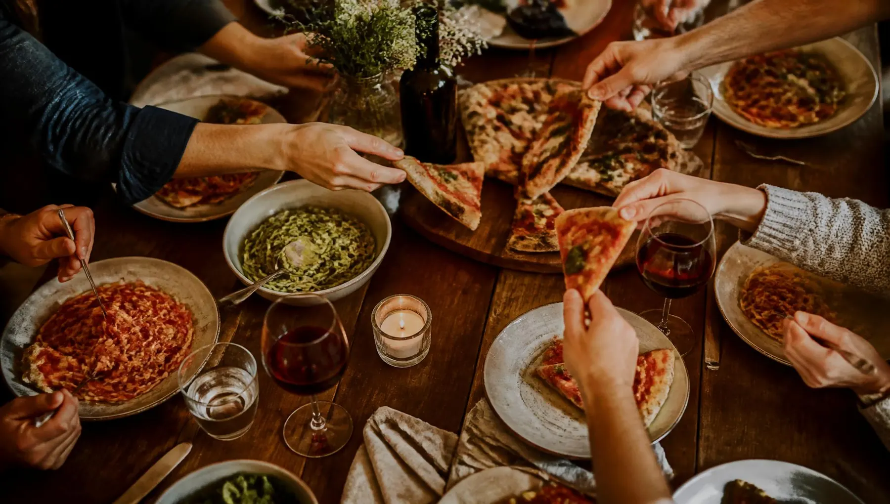Hands sharing pizza and pasta across a warm wooden table